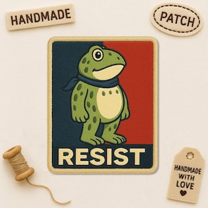 May include: Embroidered patch featuring a cartoon frog with a blue scarf, set against a red and navy background. The word "RESIST" is at the bottom. The patch is surrounded by a gold border. Additional tags read "HANDMADE" and "PATCH".