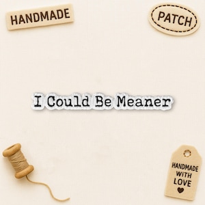 May include: A collection of craft items, featuring a fabric patch with the text "I Could Be Meaner." Other elements include a wooden thread spool, a "HANDMADE" label, and a "PATCH" tag. The background is a neutral, textured fabric.