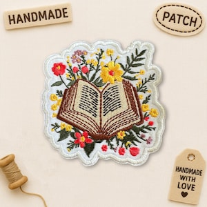 May include: Embroidered patch featuring an open book surrounded by colorful flowers. The book has detailed stitching, and the patch has a white scalloped edge. The word "HANDMADE" is on a wooden tag. The word "PATCH" is on a separate tag.