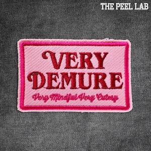 May include: A pink rectangular patch with a dark pink border and embroidered text. The text reads "VERY DEMURE" in a stylized font, with "Very Mindful Very Cutesy" below. The patch is on a dark gray fabric background.