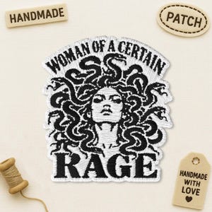 May include: A black and white embroidered patch featuring a central face surrounded by a mass of snakes, with the text "WOMAN OF A CERTAIN RAGE" arched above and "RAGE" below. The design is rendered in detailed black thread on a white fabric background.