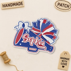 Peut inclure: Écusson brodé avec un motif de mégaphone de pom-pom girl bleu et rouge, le nom "Sophia" en cursive et un pompon. L'écusson a une bordure blanche. Une étiquette en bois indique "HANDMADE".