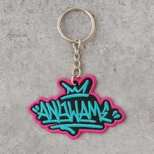 May include: A keychain with a black and teal embroidered patch. The patch has the text "AN1WAM" in a graffiti style, outlined in teal and black, with a pink border. The keychain has a silver metal ring and chain.