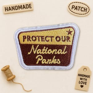 May include: Embroidered patch with the text "PROTECT OUR National Parks" in gold and brown, featuring a star. Additional tags read "HANDMADE", "PATCH", and "HANDMADE WITH LOVE". A spool of thread is also present.