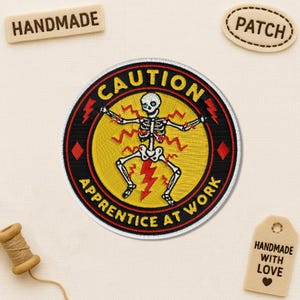 May include: Embroidered patch with a skeleton design, featuring the text "CAUTION APPRENTICE AT WORK". The patch is round with a yellow centre, red accents, and black border. Additional tags say "HANDMADE" and "PATCH".