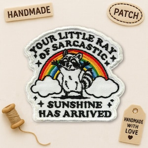 May include: Embroidered patch featuring a raccoon standing in front of a rainbow. The patch reads "YOUR LITTLE RAY OF SARCASTIC SUNSHINE HAS ARRIVED" in black text. Additional wooden tags say "HANDMADE" and "PATCH".