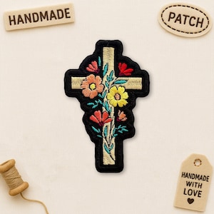 May include: Embroidered patch in the shape of a cross, featuring a floral design with orange, yellow, and red flowers. The cross has a black border and a beige background. The word "HANDMADE" is on a wooden tag.