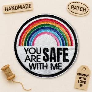 Peut inclure: Écusson brodé rond avec un arc-en-ciel et l'inscription "YOU ARE SAFE WITH ME". Le patch a une bordure noire. Des étiquettes supplémentaires indiquent "HANDMADE", "PATCH" et "HANDMADE WITH LOVE".