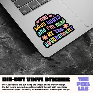 May include: A die-cut vinyl sticker with the text "my body hurts and I'm mad at The Government" in a colorful, bubble-style font. The sticker features hearts and flower accents, and is displayed on a laptop.