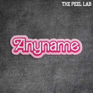 May include: A pink and white embroidered patch featuring the word "Anyname" in a cursive style. The patch has a white border and is set against a dark grey denim background. The text "THE PEEL LAB" is at the top.
