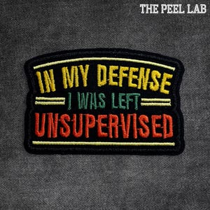 May include: Black embroidered patch with the text "IN MY DEFENSE I WAS LEFT UNSUPERVISED" in yellow, green, and red. The patch has a black border and is set against a grey background. The text "THE PEEL LAB" is in the top right corner.