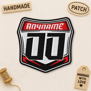 May include: Embroidered patch with a black and white design, featuring the text "ANYNAME" in red and the numbers "00". The patch is bordered in black and red, with a white background. The word "HANDMADE" is on a wooden tag.