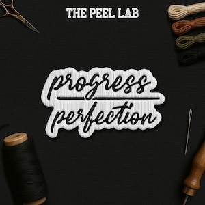 May include: A black and white embroidered patch with the words "progress" over "perfection". The patch is surrounded by sewing tools, including scissors, thread spools, and a needle, all set against a black background. The text "THE PEEL LAB" is at the top.