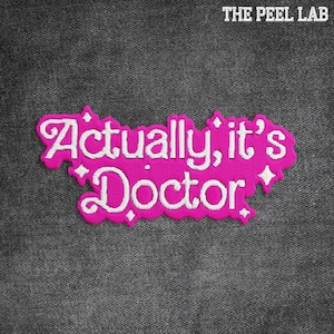 May include: A pink embroidered patch with the words "Actually, it's Doctor" in white script. The patch has a decorative border and is set against a dark gray textured background. The Peel Lab logo is in the top right corner.