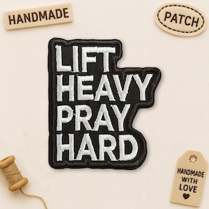 May include: Black embroidered patch with the words "LIFT HEAVY PRAY HARD" in white. The patch is surrounded by a black border. Additional tags read "HANDMADE", "PATCH", and "HANDMADE WITH LOVE". A spool of thread is also visible.