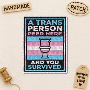 May include: Embroidered patch with the text "A TRANS PERSON PEED HERE" above a toilet graphic and "AND YOU SURVIVED" below. The patch features the transgender pride flag colors: pink, blue, and white. Handmade tag and a spool of thread are also visible.