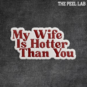 May include: Embroidered patch with the text "My Wife Is Hotter Than You" in red thread on a white background. The patch is set against a dark grey denim fabric. The Peel Lab logo is in the top right corner.