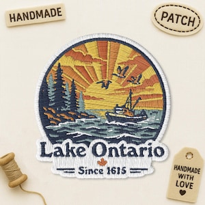 May include: A circular embroidered patch featuring a landscape of Lake Ontario with a boat on the water, pine trees on the shore, and a sunset sky with birds. The text reads Lake Ontario Since 1615 with a small orange maple leaf icon.
