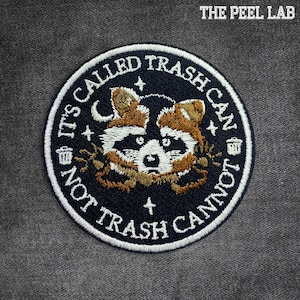 May include: Embroidered patch with a raccoon design. The patch is circular, with a dark blue background and white border. Text around the raccoon reads "IT'S CALLED TRASH CAN NOT TRASH CANNOT". The raccoon has brown and white fur. The text "THE PEEL LAB" is at the top.
