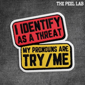 Peut inclure: Deux écussons brodés avec des bordures blanches. Le patch supérieur est rouge avec le texte "I IDENTIFY AS A THREAT" en noir. Le patch inférieur est jaune avec le texte "MY PRONOUNS ARE TRY/ME" en noir. Les écussons sont sur un tissu gris.
