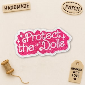May include: A pink embroidered patch with the words "Protect the Dolls" in white script. The patch has a white border and is surrounded by small star-like embellishments. Additional tags read "Handmade" and "Patch".