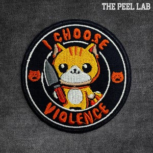 May include: Embroidered patch featuring a cartoon cat holding a knife. The patch is round with a black background and a white border. Text on the patch reads "I CHOOSE VIOLENCE" in orange. The Peel Lab logo is at the top.