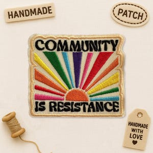 May include: Embroidered patch with the text "COMMUNITY IS RESISTANCE" in black, with a colorful sunburst design. Additional tags read "HANDMADE", "PATCH", and "HANDMADE WITH LOVE". A spool of thread is also present.
