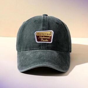 May include: A dark green baseball cap with a curved brim. The cap features a rectangular patch with the text "PROTECT OUR NATIONAL PARKS" in a vintage style. The patch has a brown border and a gold background.