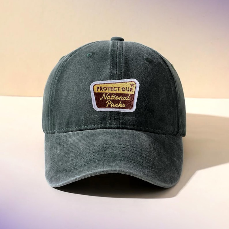 May include: A dark green baseball cap with a curved brim. The cap features a rectangular patch with the text "PROTECT OUR NATIONAL PARKS" in a vintage style. The patch has a brown border and a gold background.
