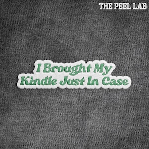May include: Embroidered patch with the text "I Brought My Kindle Just In Case" in green thread on a white background. The patch is on a dark gray denim fabric. The Peel Lab logo is in the upper right corner.