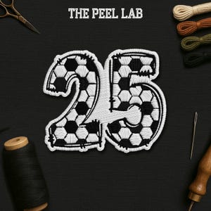 May include: Embroidered patch of the number 25, designed with a soccer ball pattern in black and white. The text "THE PEEL LAB" is at the top. The patch is surrounded by sewing supplies, including thread, scissors, and a needle.