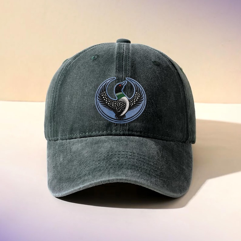 May include: A charcoal gray baseball cap with a distressed finish. Centered on the front is an embroidered circular patch featuring a stylized black and white loon with a green neck band, set against a light blue background with a crescent design.
