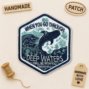 May include: A navy blue and white embroidered patch featuring the text "WHEN YOU GO THROUGH DEEP WATERS I WILL BE WITH YOU ISAIAH 43:2" and a whale design. The patch has a white border. Handmade wooden tags and a spool of thread are also visible.