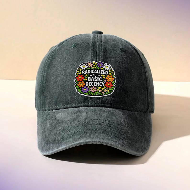 May include: A dark grey baseball cap with a curved peak. The cap has a colourful floral patch with the text "RADICALIZED by BASIC DECENCY" in a circular design. The hat is made of a soft, textured fabric.