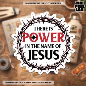 May include: A white die-cut sticker with the text "THERE IS POWER IN THE NAME OF JESUS" in black and red, surrounded by a black crown of thorns. The sticker is labeled "WATERPROOF DIE-CUT STICKERS" and "CUSTOM PORTRAITS & PLAYFUL, POSITIVE STICKER ART".
