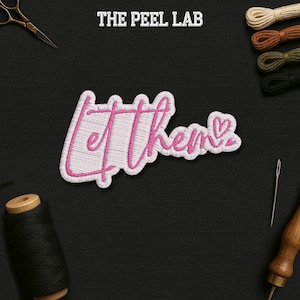 May include: Embroidered patch with the words "Let Them" in pink script, with a heart over the "s". The patch is white with pink stitching. The text "THE PEEL LAB" is at the top. Sewing supplies are in the background.