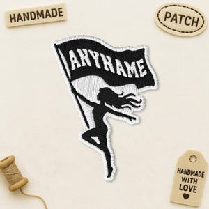 May include: An embroidered patch featuring a black silhouette of a person with long hair holding a flag. The flag is black with white borders and contains the white text ANYNAME. The patch has a white background and white stitched edges.