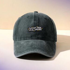 May include: A dark green baseball cap with a curved brim. The cap features the embroidered text "SUPPORT YOUR LOCAL LIBRARY" in white and yellow. The hat is made of a soft, worn material.