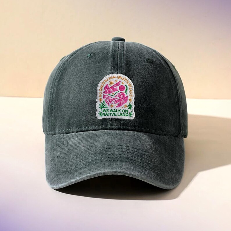 May include: A dark green baseball cap with a curved brim. The cap features a patch with the text "NO ONE IS ILLEGAL ON STOLEN LAND" and "WE WALK ON NATIVE LAND" with a pink and white graphic.