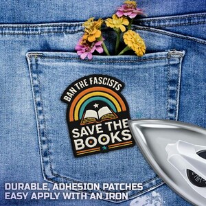 May include: A black and colorful patch with the text "BAN THE FASCISTS" and "SAVE THE BOOKS" is placed on a blue denim pocket. The patch features a rainbow and an open book design. A few colorful flowers are in the pocket. An iron is in the image.