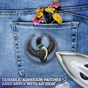 May include: A circular embroidered patch featuring a black and white loon with a green neck band and red eye, set against a blue denim pocket with yellow and pink flowers. A silver iron is positioned in the lower right corner of the image.