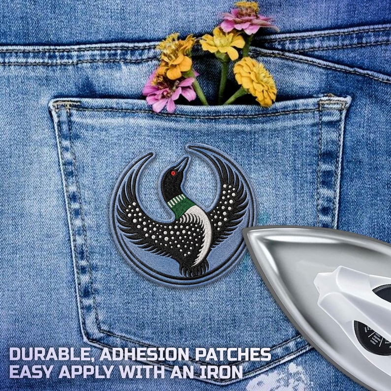 May include: A circular embroidered patch featuring a black and white loon with a green neck band and red eye, set against a blue denim pocket with yellow and pink flowers. A silver iron is positioned in the lower right corner of the image.