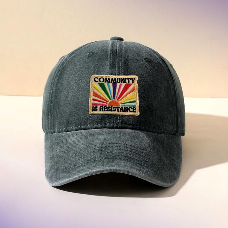 May include: A dark teal baseball cap with a curved brim. The cap features an embroidered patch with the words "COMMUNITY IS RESISTANCE" above a rainbow sun design. The hat is made of a soft, worn fabric.