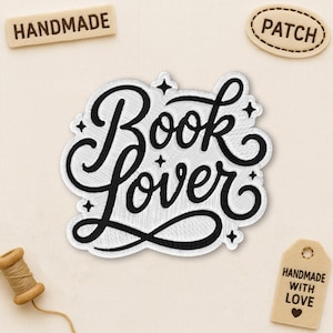 May include: Embroidered patch with the words "Book Lover" in a stylish, cursive font. The patch is white with black stitching and a black border. Additional tags read "Handmade" and "Patch". A small spool of thread and a tag that says "Handmade with Love" are also present.
