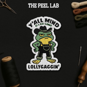 May include: Embroidered patch featuring a grumpy green frog wearing a cowboy hat and overalls. The patch reads "Y'all mind if I do some lollygaggin'?" in white thread. The Peel Lab logo is at the top. The patch is on a black background.
