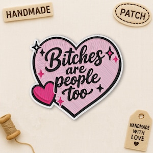 Peut inclure: Écusson brodé rose en forme de cœur avec l'inscription "Bitches are people too" en script noir. Un petit cœur rose est attaché sur le côté gauche. D'autres écussons et une bobine de fil sont également visibles.