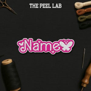 May include: Embroidered patch with the word "Name" in pink and white, featuring a butterfly design. The patch is set against a black background with sewing tools and thread spools. The text "THE PEEL LAB" is at the top.