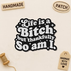 May include: Embroidered patch with the phrase "Life is a Bitch but thankfully So am I." in white lettering on a black background. The patch is surrounded by white stars. Additional tags read "Handmade" and "Patch".