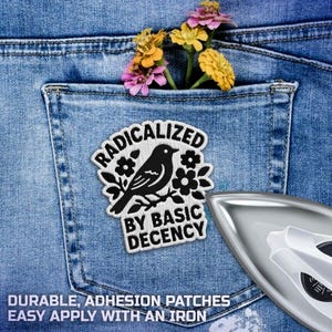 Pode incluir: Um bolso de ganga azul com um patch com as palavras "RADICALIZED BY BASIC DECENCY" e um desenho de p&aacute;ssaro preto. Flores coloridas est&atilde;o presas no bolso. Um ferro de engomar est&aacute; no canto inferior direito.