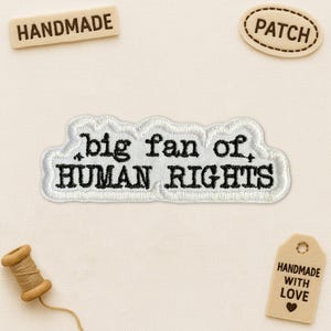 May include: Embroidered patch with the text "big fan of, HUMAN RIGHTS" in black on a white cloud-shaped background. The image also includes wooden tags that say "HANDMADE", "PATCH", and "HANDMADE WITH LOVE". A spool of thread is also visible.